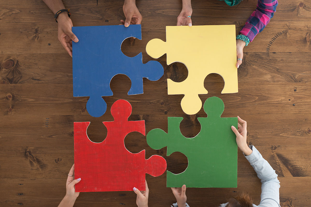 Vier Menschen stehen um einen Tisch und halten je ein Puzzelteil aneinander.