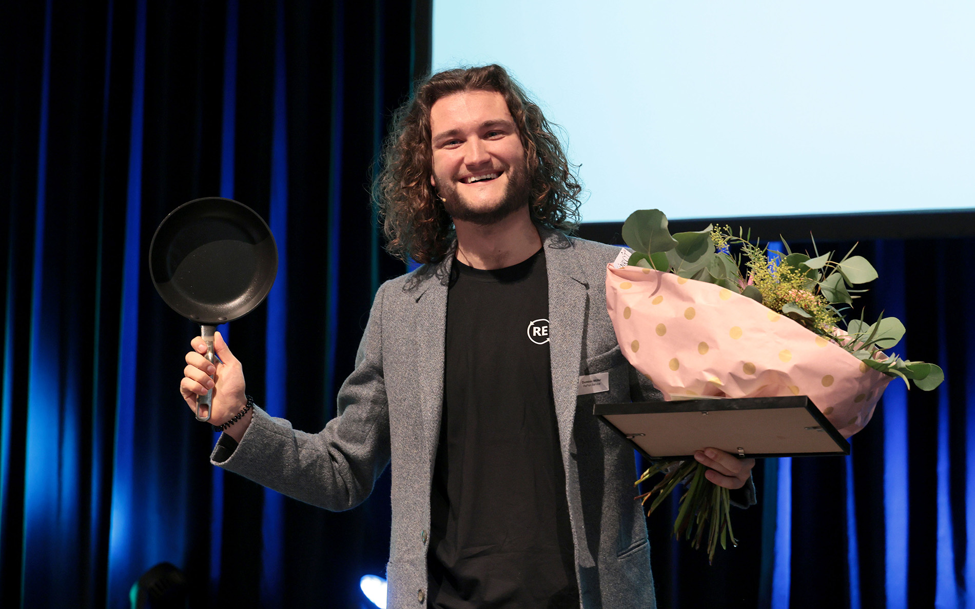 Dominik Müller steht auf der Bühne und lacht. Er hält in einer Hand eine kleine Pfanne und in der anderen einen Blumenstrauss.